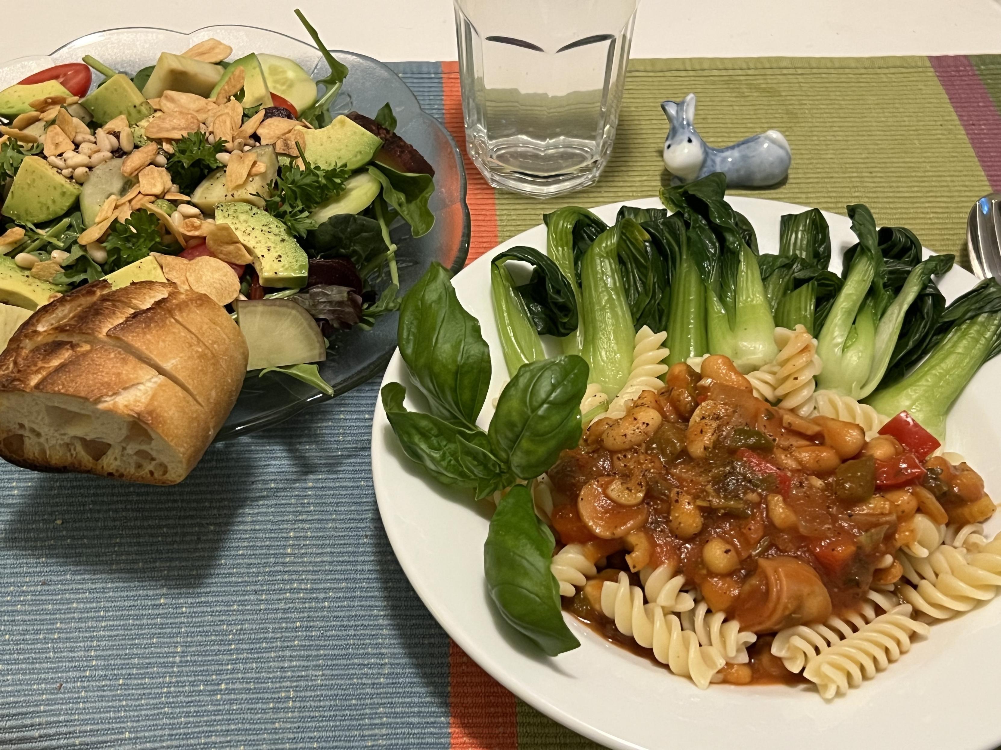 A big dinner bowl of rotini pasta with veggie tomato sauce with northern white beans, red bell pepper and eryngii mushroom, garnished with basil leaves. Boiled baby bok choys are also  on the wide rim of the same bowl.
On another bowl equally big as the pasta one has green leafy greens and arugulas topped with slices of cooked beet, satsuma sweet potato, fresh avocado, cucumber, pine nuts, and garlic crunch. No oil, just rice vinegar and black pepper to taste. There’s also a piece of toasted baguette on the rim of the salad bowl. 
A glass of lemon water and a bunny hashi-oki are also on the placemat.